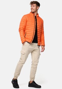 Orange polstret jakke med ståkrave, sort lynlås, båret over en sort skjorte, parret med lyse beige cargo-bukser og sorte sneakers.