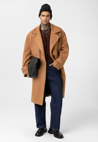 Man met nektattoo, draagt zwarte beanie, camel-kleurige overjas, donkere trui, blauwe jeans, zwarte loafers, houdt een zwart opgevouwen tas vast voor een witte achtergrond.