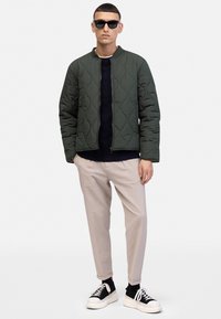 Grüne gesteppte Bomberjacke mit Reißverschluss, getragen über einem marineblauen Pullover, kombiniert mit hellbeigen, schmal zulaufenden Hosen und schwarzen Sneakern.