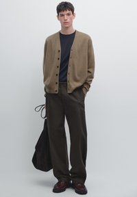 Beige cardigan with buttons over a navy shirt, paired with dark brown trousers. Model holds a dark bag and wears brown shoes.