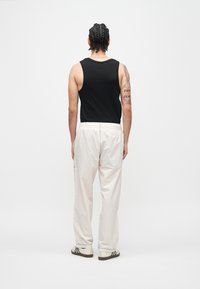 Μαύρο αμάνικο πουκάμισο, λευκό παντελόνι με ελαστική μέση, πίσω όψη που δείχνει πλεγμένα μαλλιά και τατουάζ. Τα παπούτσια έχουν τρεις μαύρες ρίγες.