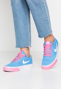 Blå canvas sneakers med rosa snören, vit swoosh-logotyp och rosa gummitätning. Har en låg design och texturerad yta för grepp.