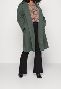 Abrigo largo de mezcla de lana verde con un corte relajado, que presenta un cuello con muescas y bolsillos laterales, superpuesto a una blusa estampada y pantalones negros acampanados.