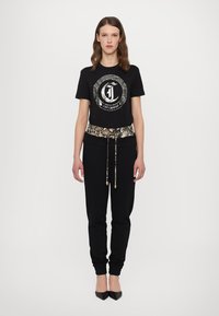 Svart T-skjorte med sirkulært logo, kombinert med svarte joggebukser som har et belte med slanger mønster. Modellen har på seg spisse svarte hæler.