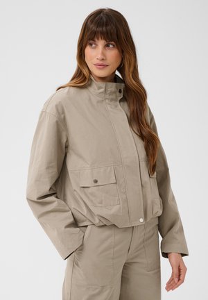 Donna con capelli lunghi e castani che indossa una giacca beige a collo alto con tasche anteriori e pantaloni abbinati su uno sfondo chiaro uniforme.