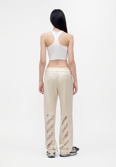 Femme aux longs cheveux noirs portant un crop top blanc dos nageur, un pantalon crème avec des découpes en dentelle et des baskets blanches, debout de dos sur un fond blanc.