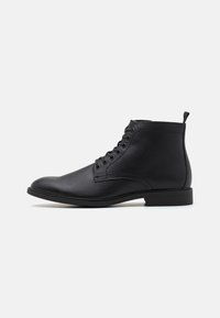 Botas de cuero negras hasta el tobillo con punta redonda, cierre de cordones en la parte delantera y suela baja de goma. Incluye una lengüeta en la parte trasera para facilitar su colocación.
