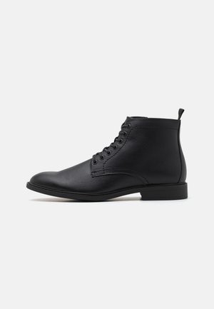 Schwarze Lederstiefeletten mit runder Zehenkappe, Schnürung vorn und einer flachen Gummisohle. Mit einer Zuglasche auf der Rückseite für einfaches Anziehen.