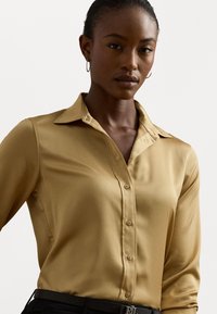 Blusa a maniche lunghe di colore marrone chiaro, realizzata in un tessuto liscio, con colletto appuntito e chiusura a bottoni sul davanti. Cintura nera visibile in vita.