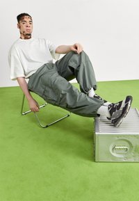 Schwarze und graue Sneaker, grüne Cargo-Hose und ein oversized weißes T-Shirt. Modell sitzt auf einem Chromstuhl mit einer metallenen Kiste, vor einem grünen Boden.