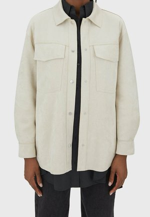 Personne portant une veste crème boutonnée avec des poches poitrine à rabat sur une chemise noire et un pantalon foncé, debout devant un fond uni.