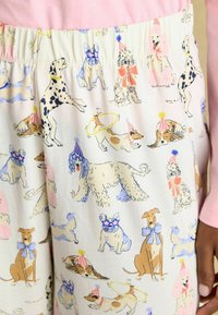 Ženske pižame z vzorcem, ki vsebujejo različne ilustracije psov v modri, rumeni in roza barvi na kremnem ozadju. Opažena je elastična pasu.