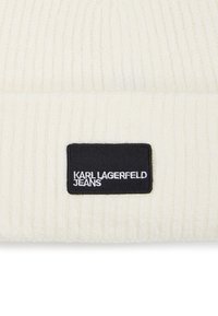 Ribbet kremfarget lue med brettet kant, prydet med en svart etikett som har teksten "KARL LAGERFELD JEANS" i hvit skrift.
