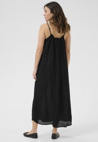 Donna con capelli castano di lunghezza spalla, che indossa un abito maxi nero senza maniche con spalline sottili e ballerine nere, in piedi contro uno sfondo grigio.