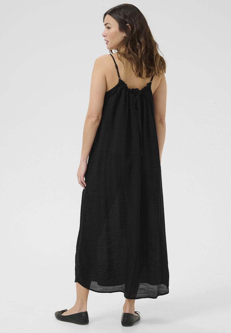 Donna con capelli castano di lunghezza spalla, che indossa un abito maxi nero senza maniche con spalline sottili e ballerine nere, in piedi contro uno sfondo grigio.