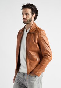 Brun læderjakke med krave, glat tekstur, frontlynlås og syninger, bæres over en lysegrå sweater og jeans.