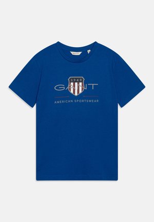 Blå bomuld T-shirt med rund halsudskæring og korte ærmer. Forsiden har et skjoldlogo med striber og teksten "GANT AMERICAN SPORTSWEAR."