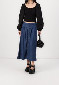 Haut noir à manches longues avec texture froncée, associé à une jupe midi en denim bleu boutonnée sur le devant. Porté avec des chaussures à plateforme noires et un sac à main imprimé.