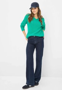 Grøn ribbet sweater, mørkeblå brede jeans og en marineblå kasket. Outfittet har en afslappet pasform, teksturerede detaljer og kontrasterende farver.