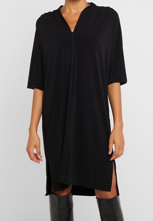 Vrouw met een losse zwarte jurk tot aan de knie, met halflange mouwen, zijsplitten en een V-hals, gecombineerd met zwarte kniehoge laarzen.