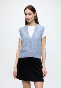 Blauwe gebreide gilet met gestructureerd garen, vijf knopen en een V-hals. Draagt over een wit T-shirt en gecombineerd met een zwarte ribfluwelen rok.