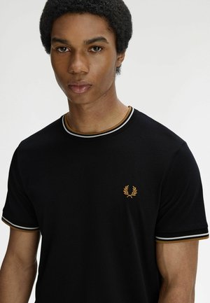 Joven con cabello corto y rizado que lleva una camiseta negra con ribetes blancos y dorados y un pequeño logo de corona de laurel dorada en el pecho.