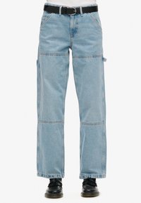 Jeans de mezclilla de corte amplio en un azul claro, con múltiples bolsillos, un corte recto y costuras visibles. Combinados con zapatos negros y un cinturón.