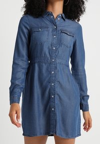 Jeansklänning med långa ärmar, ljusblått tyg, knäppning fram, två bröstfickor och insydd midja.