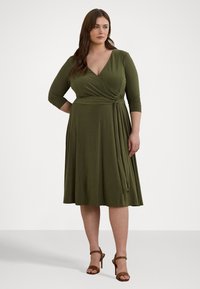 Ελιάς πράσινο, φόρεμα τύπου περιτυλίγματος με τρία τέταρτα μανίκια, λεπτομέρεια με κόμπο στο πλάι και ροή φούστας. Φορεμένο με καφέ γόβες.