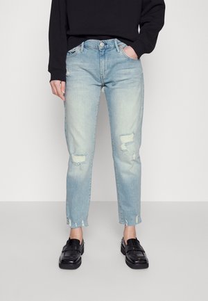 Jasnoniebieskie postrzępione dżinsy noszone z czarnymi mokasynami i czarną bluzką z długim rękawem, ręce umieszczone w przednich kieszeniach.