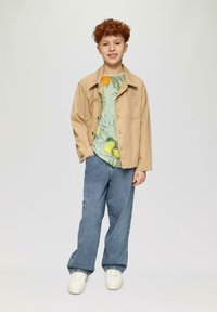 Tan, lichte jas over een lichtgroen graphic T-shirt met fruit- en bloemontwerpen; blauwe jeans; witte sneakers. Studio achtergrond.