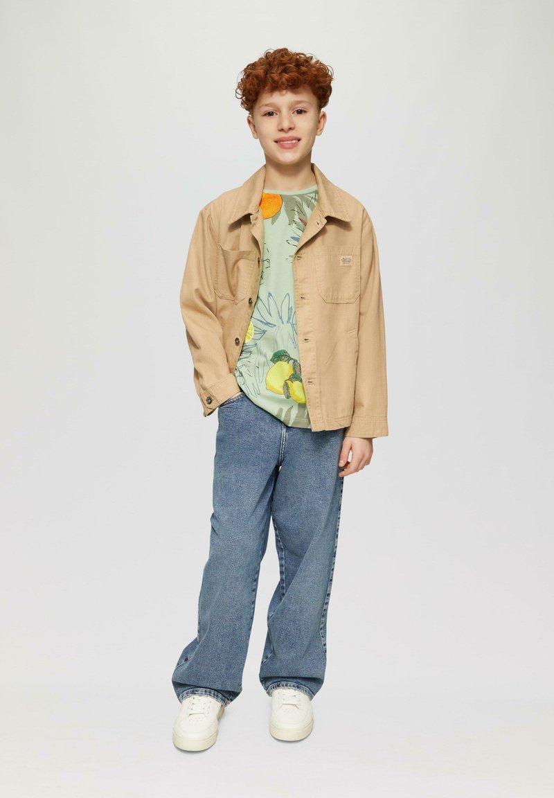 Tan, lichte jas over een lichtgroen graphic T-shirt met fruit- en bloemontwerpen; blauwe jeans; witte sneakers. Studio achtergrond.