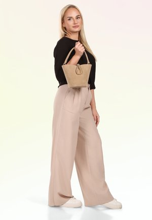 Frau in beigen, weit geschnittenen Hosen und schwarzem Oberteil, die eine hellbraune Wildleder-Bucket-Tasche mit Schlaufen-Detail hält, steht vor weißem Hintergrund.