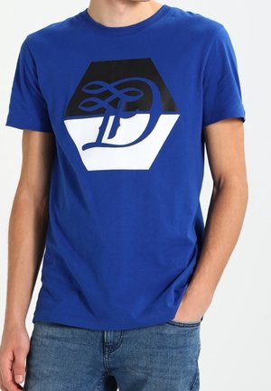 Mann trägt ein königsblaues T-Shirt mit kurzen Ärmeln und einem schwarz-weißen Hexagon-Design mit dem stilisierten Buchstaben "D", kombiniert mit blauen Jeans.