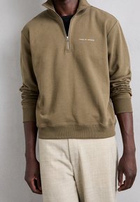 Felpa khaki con zip, polsini e orlo a coste, con logo bianco, abbinata a pantaloni beige chiaro testurizzati. Tessuto morbido.