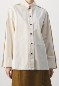Krämfärgad oversized knäppskjorta med krage, bröstficka och bruna knappar. Har lång ärmar och en avslappnad passform.