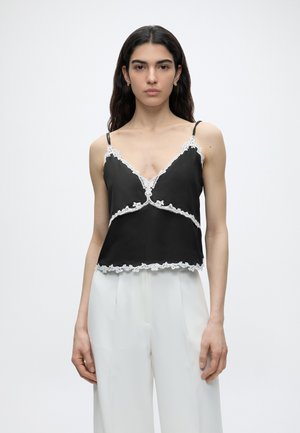 Jonge vrouw met lang donker haar draagt een zwarte cami met witte kanten rand en witte wijde broek met hoge taille, staand tegen een effen achtergrond.