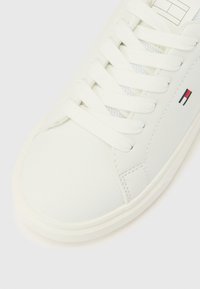 Bela superga z belimi vezalkami, teksturirano podplatom in majhno rdeče-belo-modro pravokotno oznako na strani.