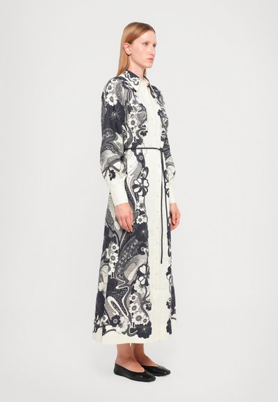 Vestido camisero largo y estampado en blanco y azul marino con diseños florales y de paisley, con botones en el frente y un lazo en la cintura. Zapatos planos negros.
