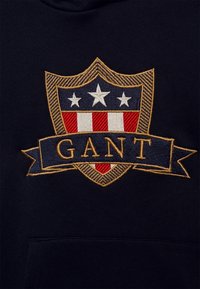 Navy hoodie featuring an embroidered shield logo with red, white, and blue accents, gold outlines, and the word "GANT" on a banner.