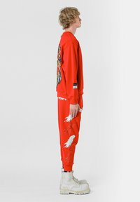 Sudadera roja con un gran gráfico colorido en la parte posterior, combinada con pantalones rojos que tienen diseños de plumas blancas. Llevada con botas de combate blancas.