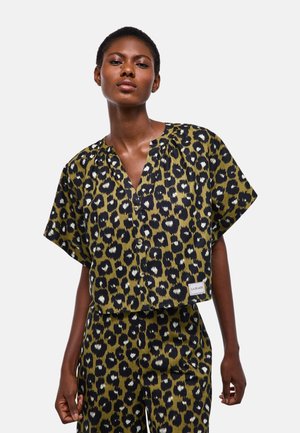 Femme portant une chemise à manches courtes et un pantalon assortis à imprimé léopard vert olive et noir, debout devant un fond blanc uni.