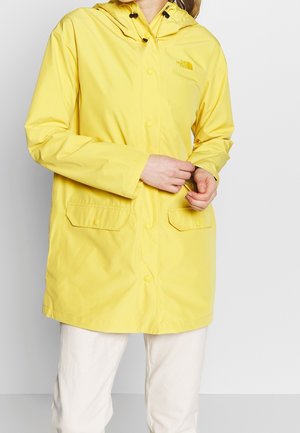 Personne portant une veste imperméable jaune vif avec capuche, boutons-pression à l'avant, poches à rabat et pantalon de couleur claire sur un fond uni.
