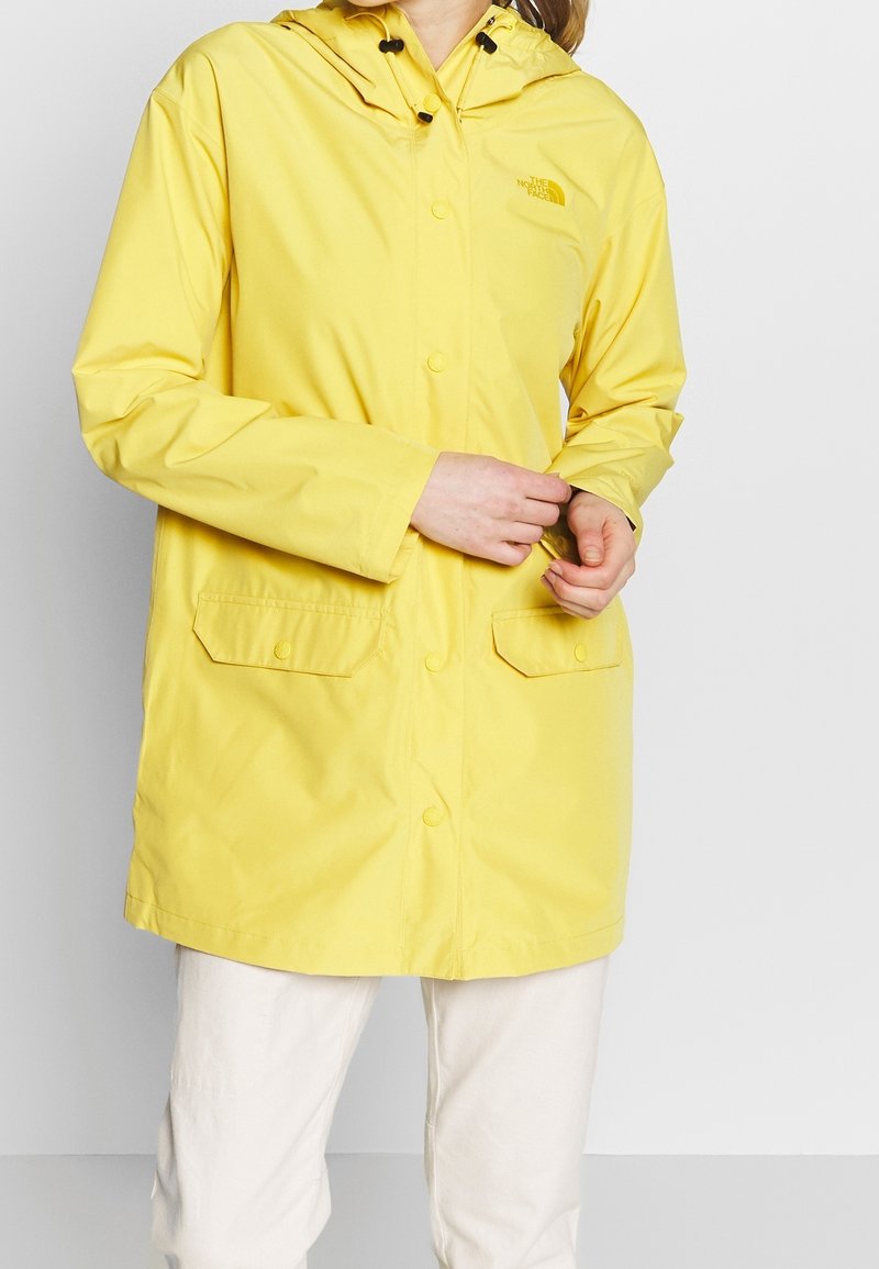 Personne portant une veste imperméable jaune vif avec capuche, boutons-pression à l'avant, poches à rabat et pantalon de couleur claire sur un fond uni.