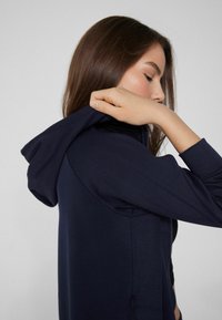 Sudadera de cremallera azul marino hecha de tela suave, con capucha y aberturas laterales. Los puños son ajustados y el diseño es minimalista con una textura suave.
