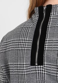 Svart- och vitrutigt zip-up-top med hög krage och en solid svart sammetstrimma längs dragkedjan för kontrast. Texturerad tyg.