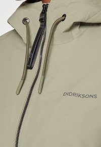 Svetle olivově zelená bunda se zipem, nastavitelnou kapucí a stahovacími šňůrkami. Obsahuje černý zip a potištěné logo na hrudi.