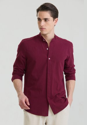 Mann trägt ein langärmliges, bordeauxrotes Hemd mit hochgekrempelten Ärmeln und hellen Hosen, steht vor einem einfarbigen Hintergrund.