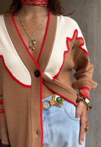 Nő barna-fehér kardigánban, piros szegéllyel, kék farmerral, barna övvel zöld kövű csattal, arany nyaklánccal charmokkal és arany karkötővel.