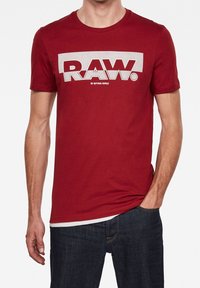 Red cotton t-shirt featuring a bold white "RAW." logo across the chest. Short sleeves and a standard crew neck. Casual fit.